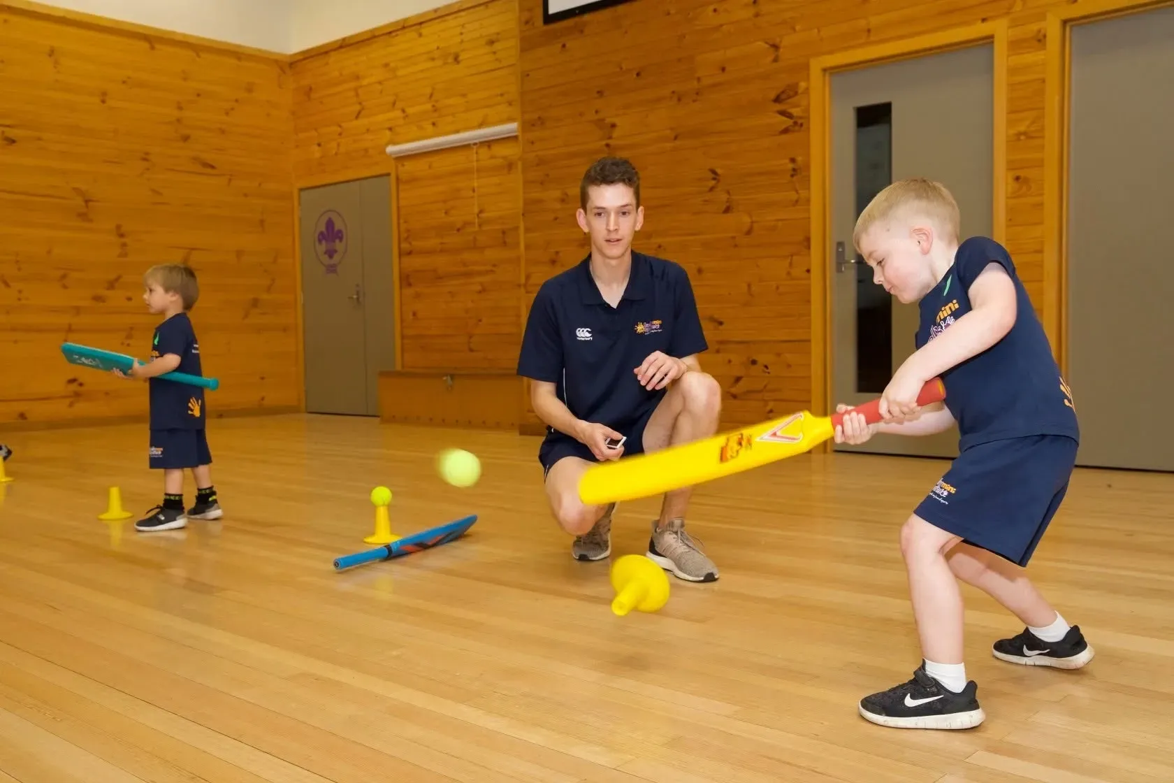 Children playing indoor hockey in a gym with a coach watching nearby