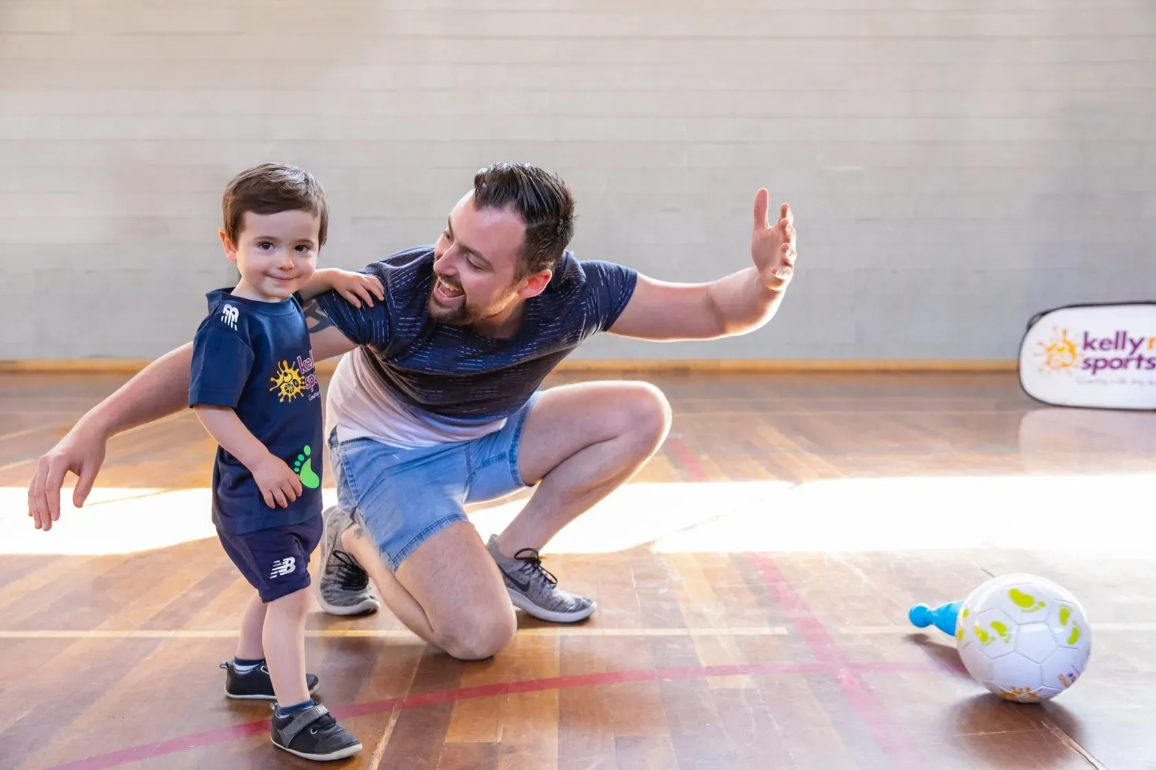 Man and child crouching on a gym floor, smiling and reaching out near a ball.