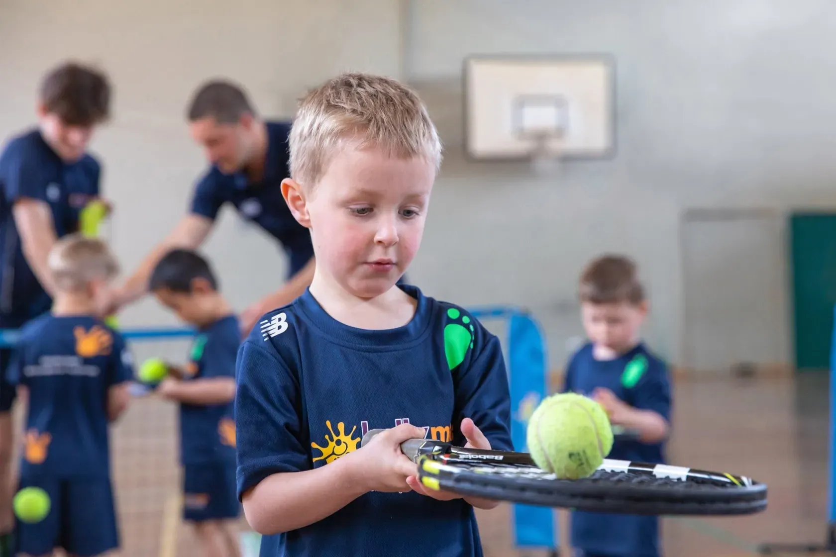 Child holding a tennis racket with a green ball in a gym, with others practicing in the background