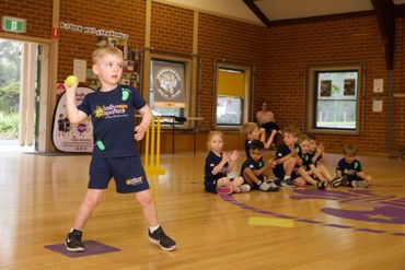 Child indoors preparing to throw a ball during a gym activity, with other children sitting nearby.