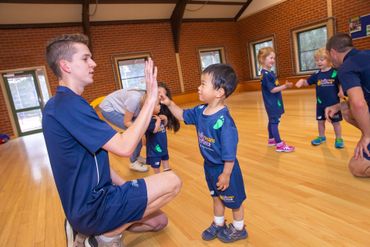 Coach kneels to high-five a child in a gym during a kids’ sports class