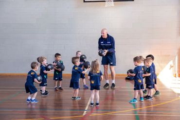 Children in navy uniforms gather around a coach in a gym, playing a group activity.
