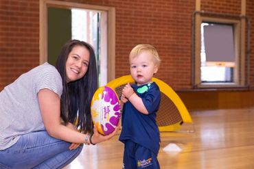 Adult and toddler holding a colorful ball in a gymnasium, with smiling adult crouched beside them