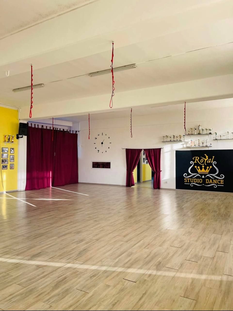 Studio di danza vuoto con pavimento in legno, tende rosse e una parete gialla. È visibile un cartello con la scritta