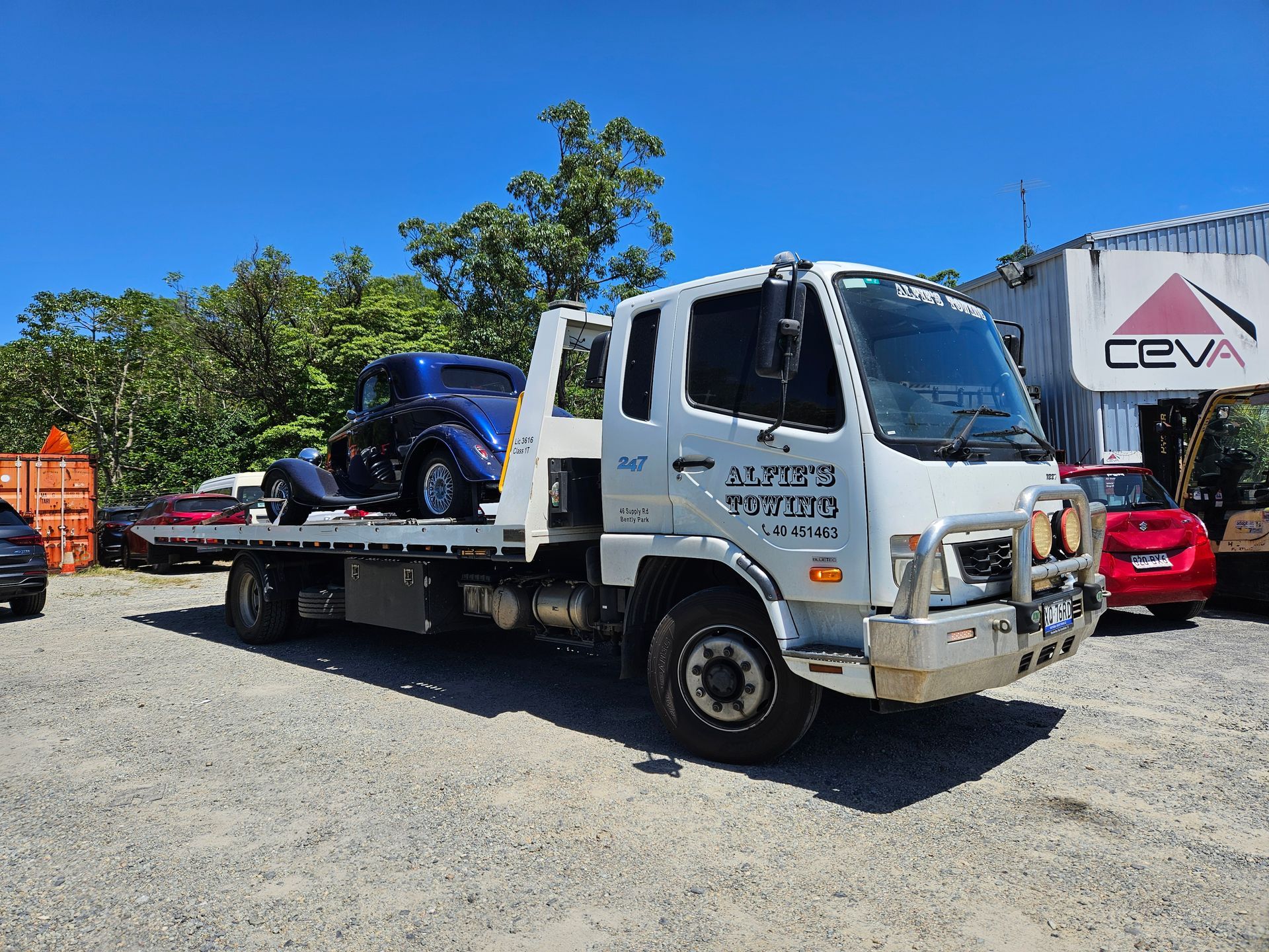 White tow truck with a blue vintage car on its flatbed, parked outside a building — Alfie's Towing in Mareeba, QLD