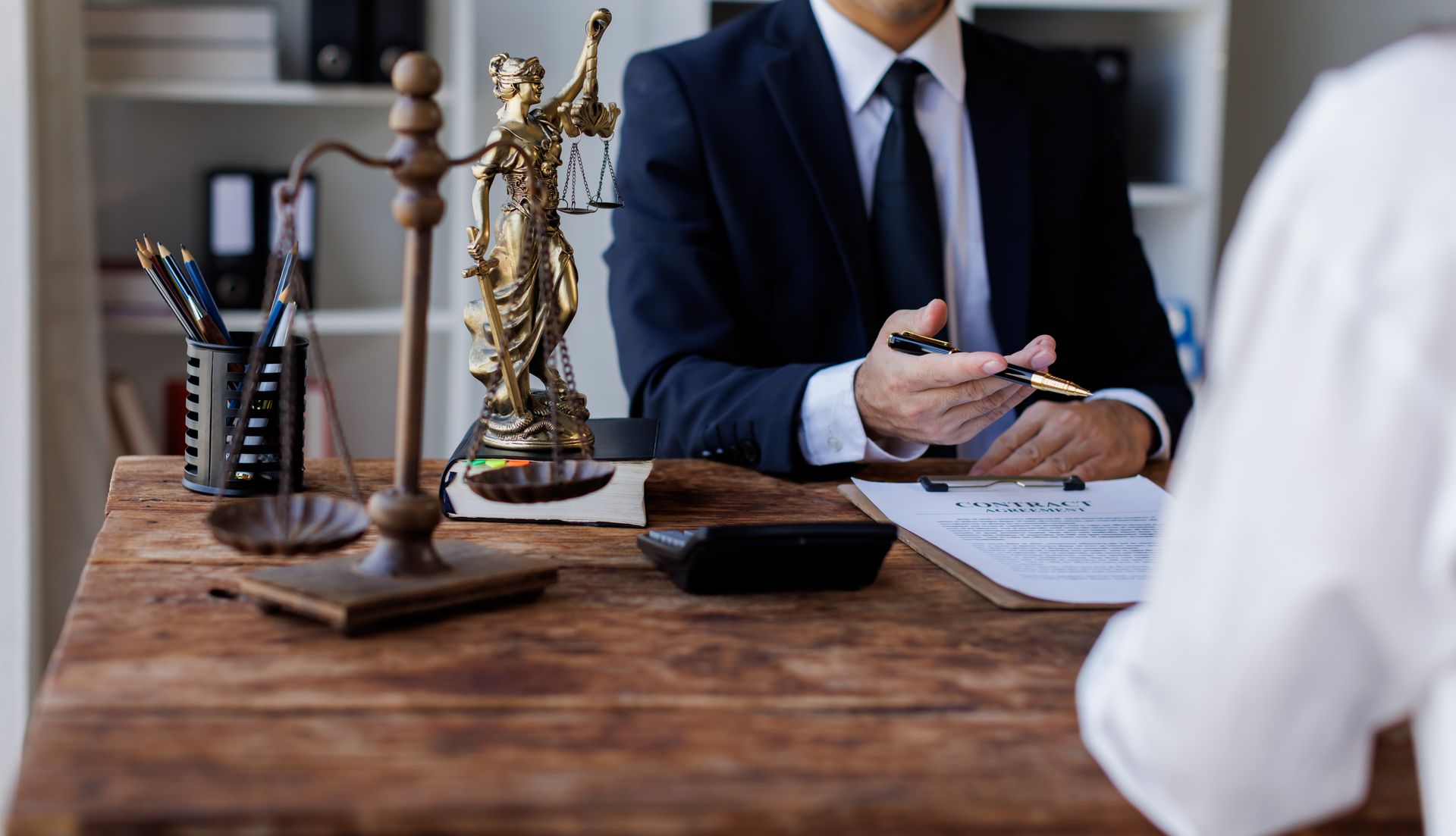 Lawyer in suit reviews document with client at wooden desk, scales of justice prominent.
