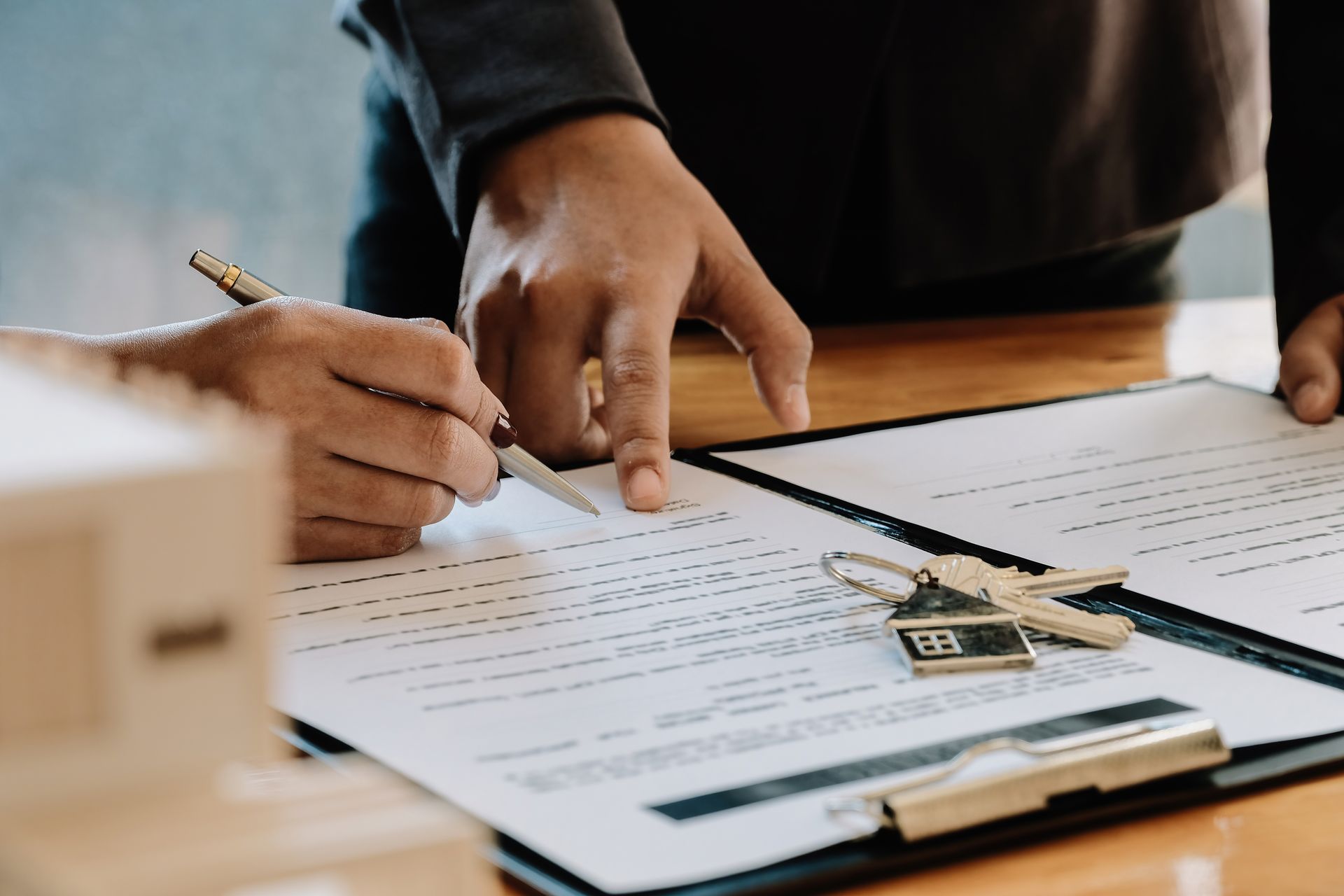 Hands signing a document, keys on top. Another hand points at the document.