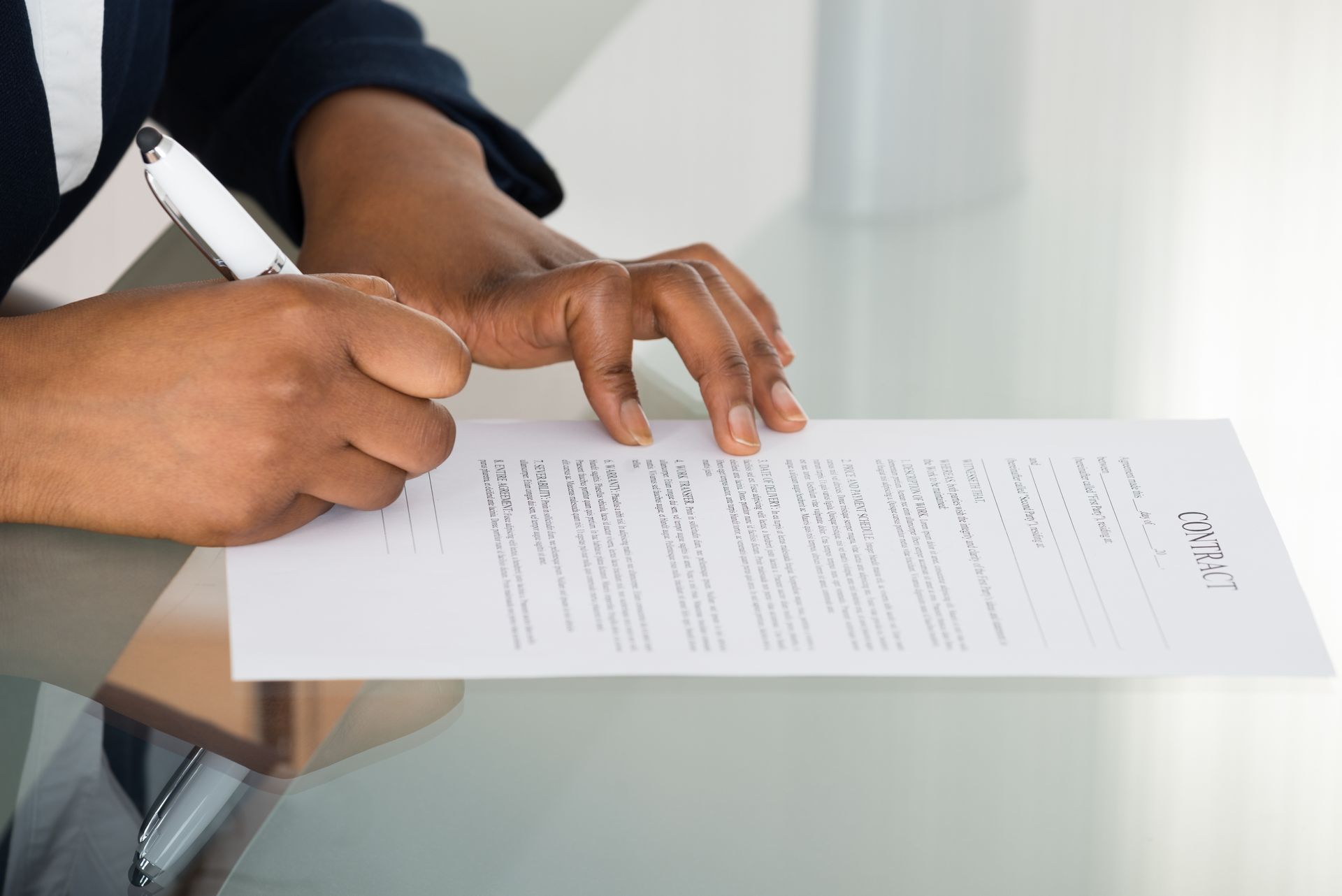 Person signing a document with a pen on a clear glass table.