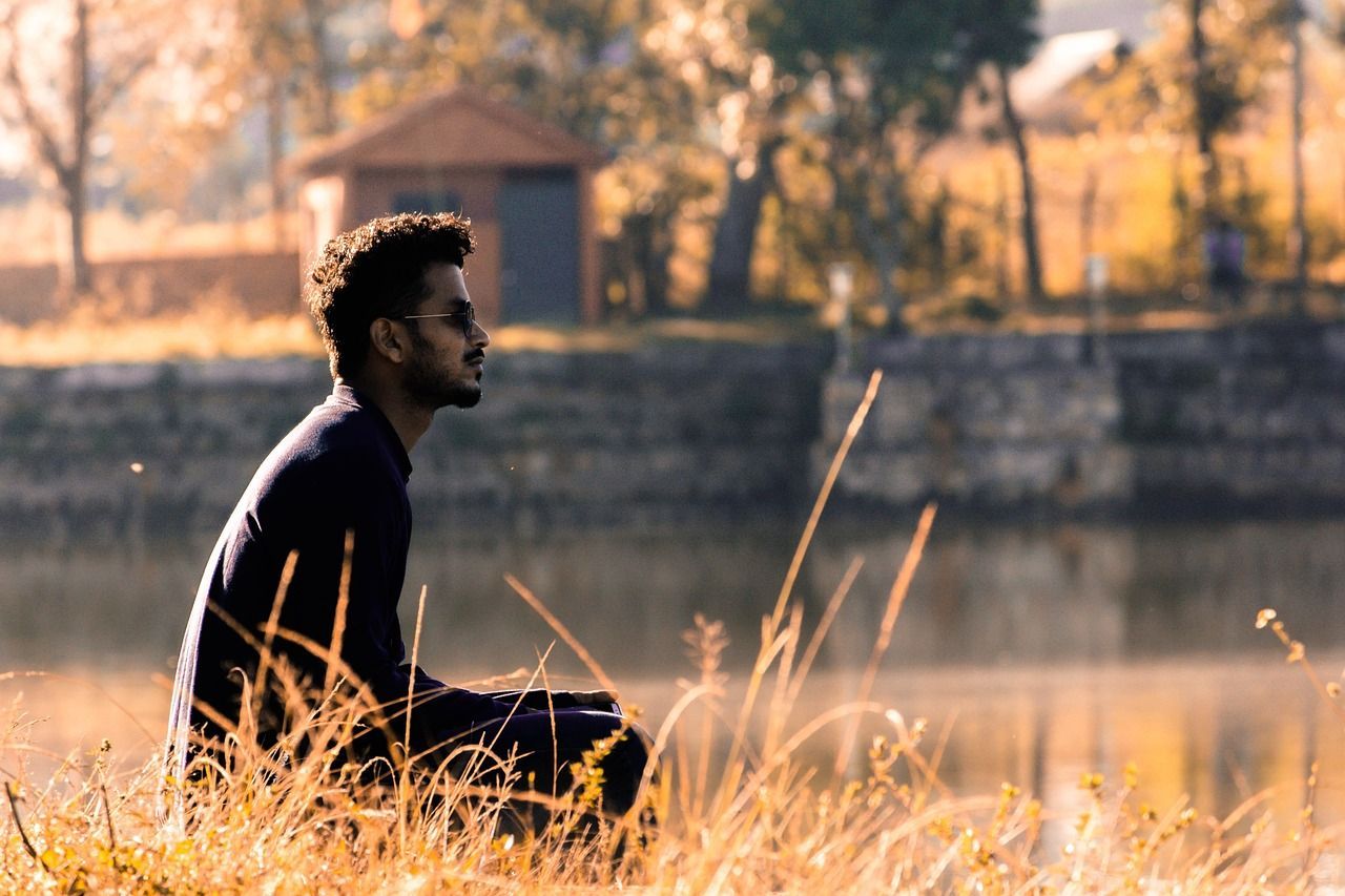Man sitting by a lake, in profile, eyes closed, near tall grass. Wooden shed and trees in the background.