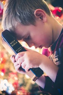 Young boy with head bowed, holding a Bible, possibly praying, against a blurred background of Christmas decorations.
