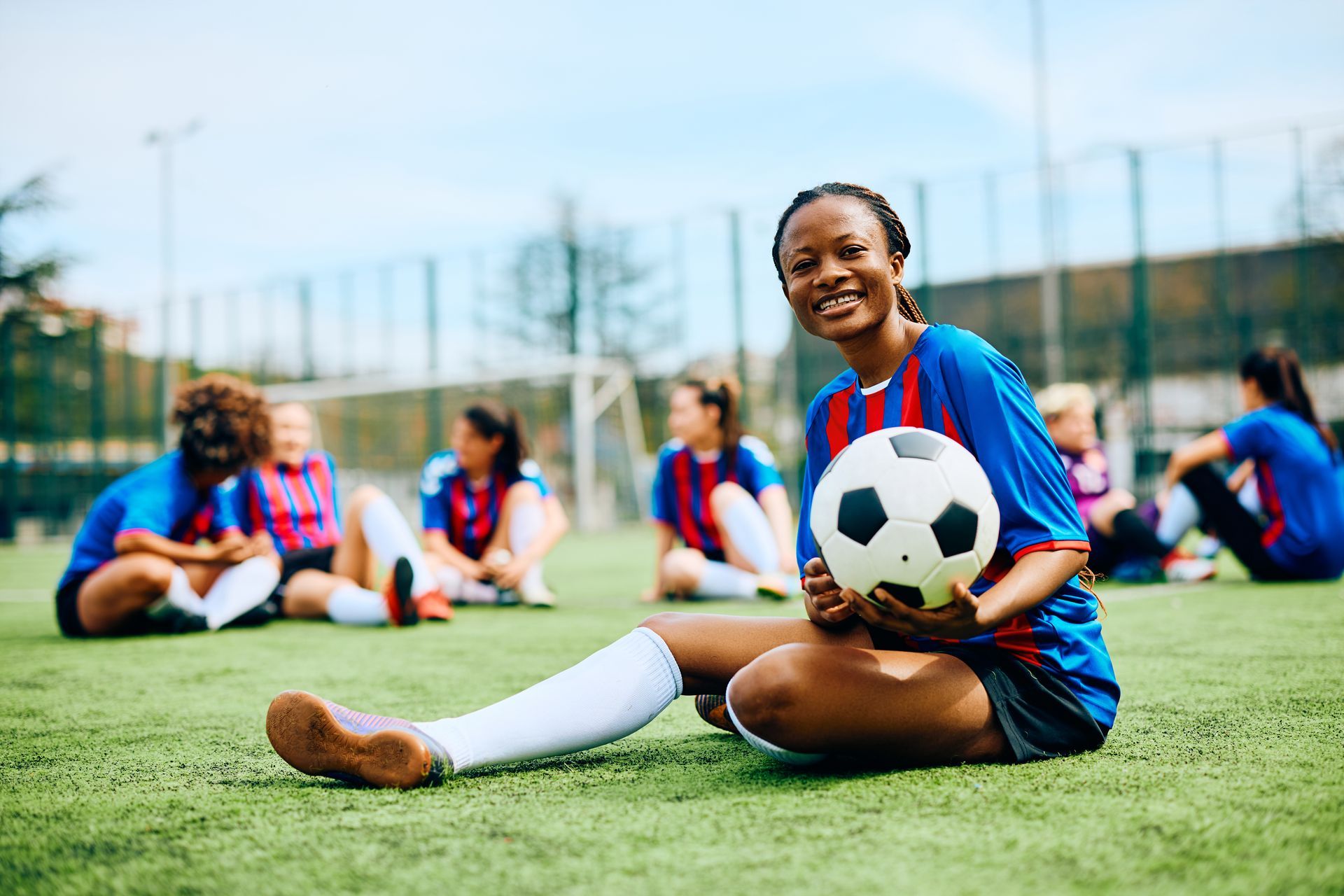 Soccer player in blue and red uniform sits on field holding a ball, with teammates blurred in the background