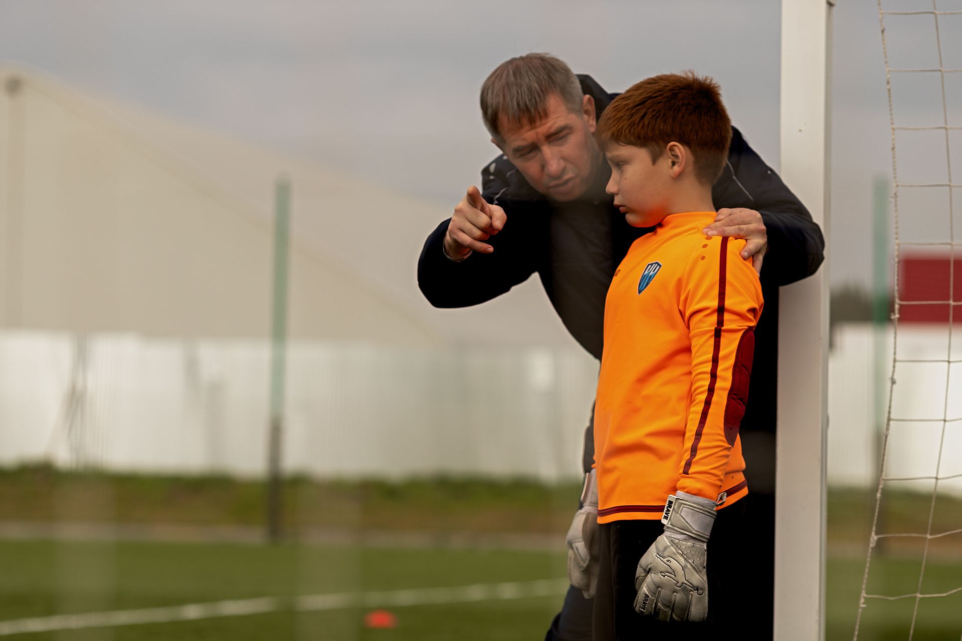 Coach speaks to a young goalkeeper in an orange jersey by the soccer goal