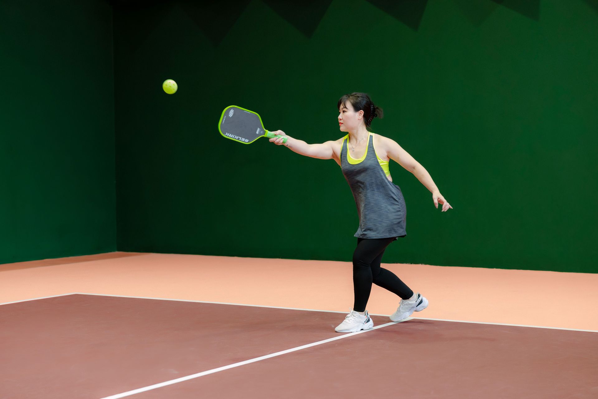 Tennis player in gray shirt hitting a ball on an indoor court with green walls and a clay-colored floor