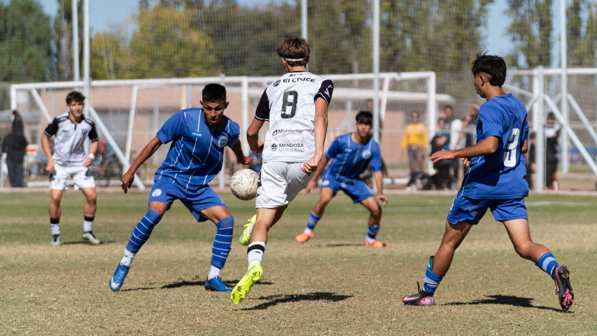 Soccer player in blue and red uniform sits on field holding a ball, with teammates blurred in the background