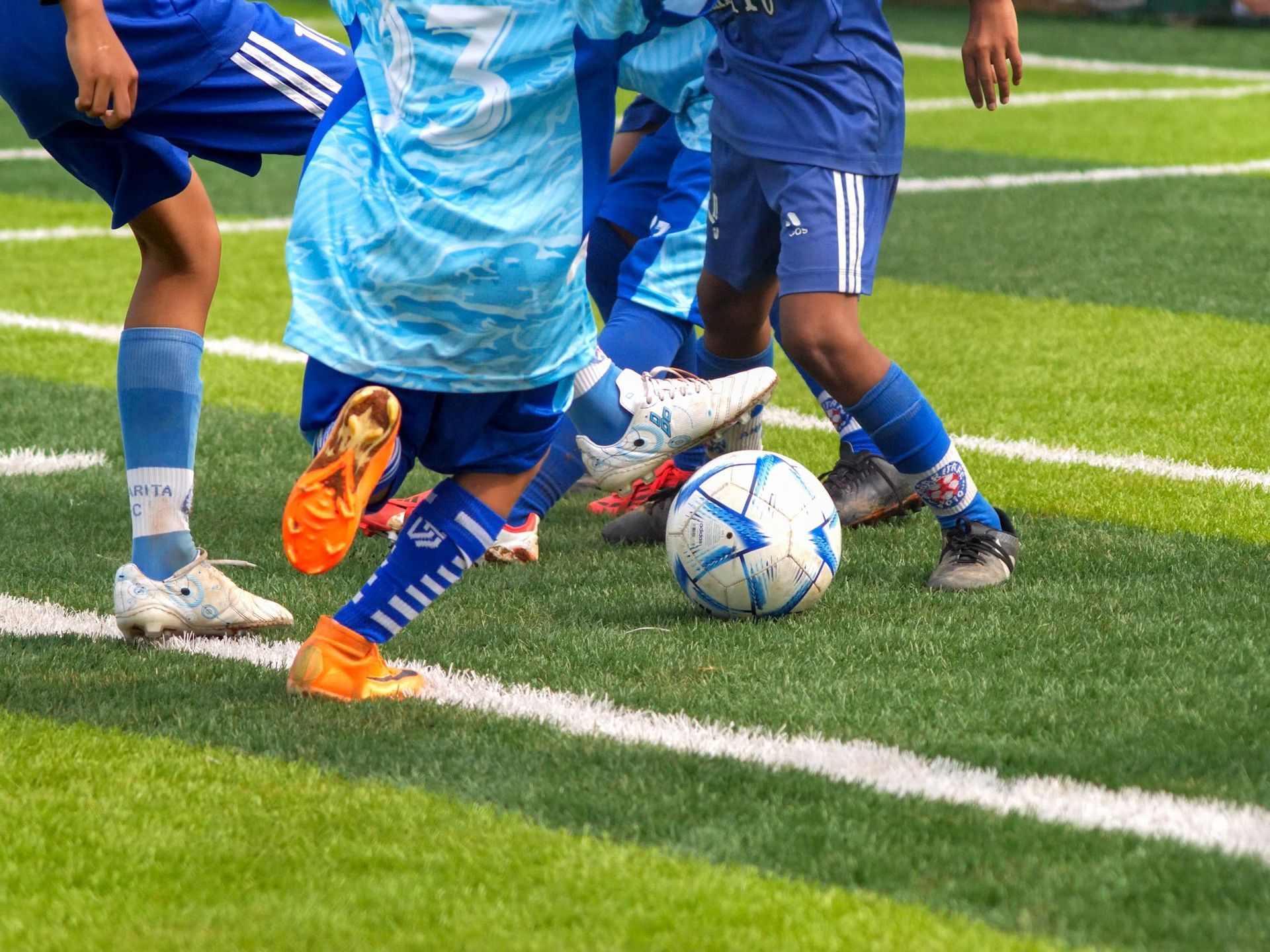 Soccer player in blue and red uniform sits on field holding a ball, with teammates blurred in the background