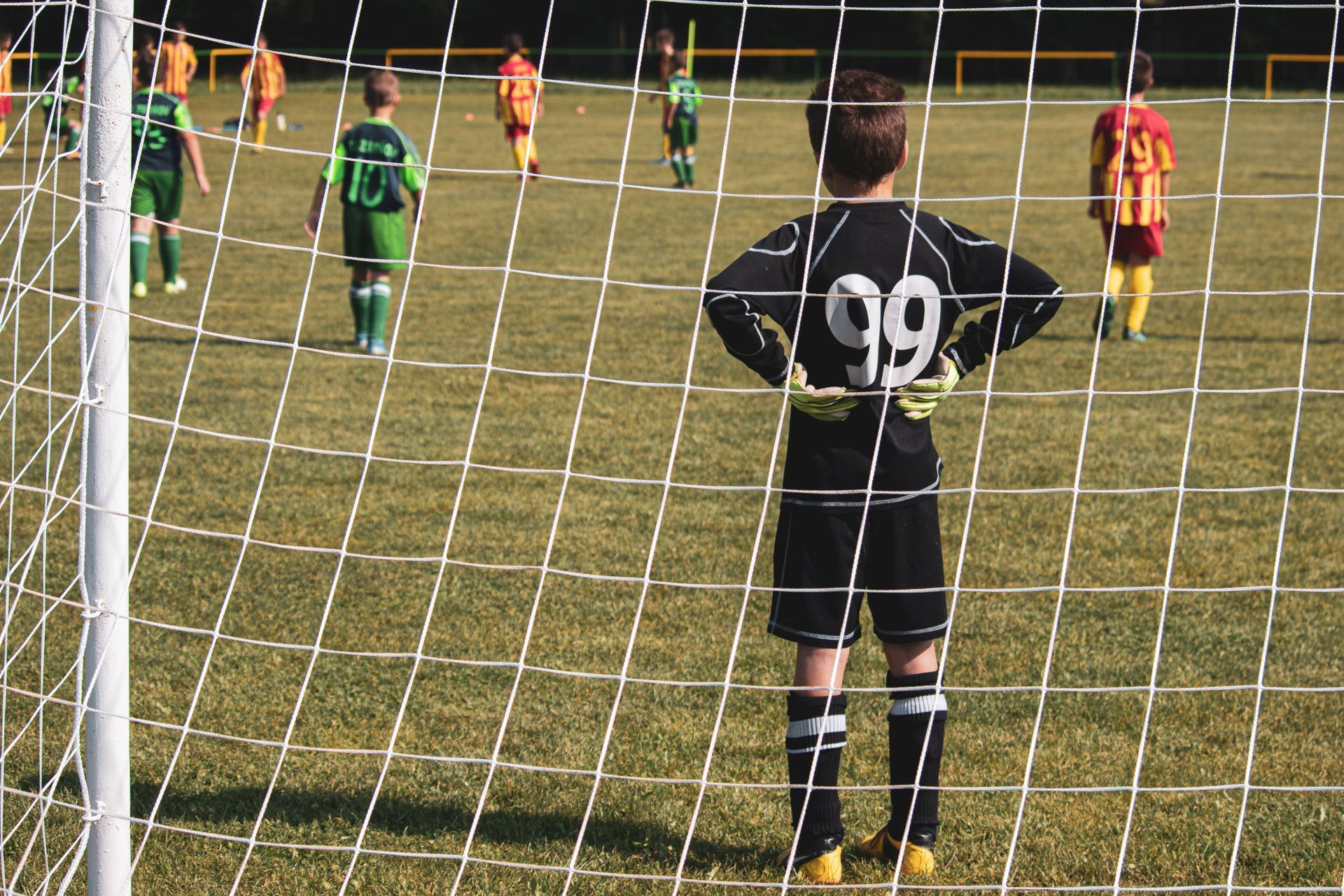 Soccer goalkeeper in black jersey number 99 stands behind the net, watching play on the field.