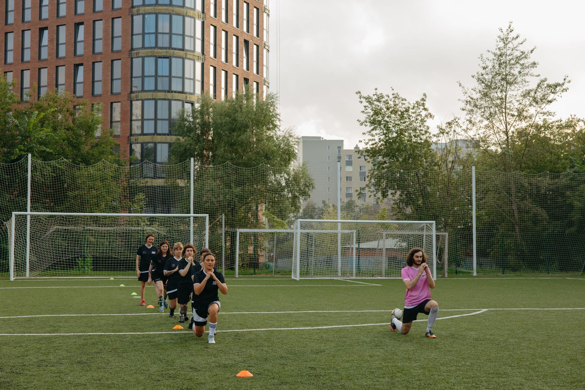 Youth soccer practice on a field, with players in black running toward a coach in pink near a goal.