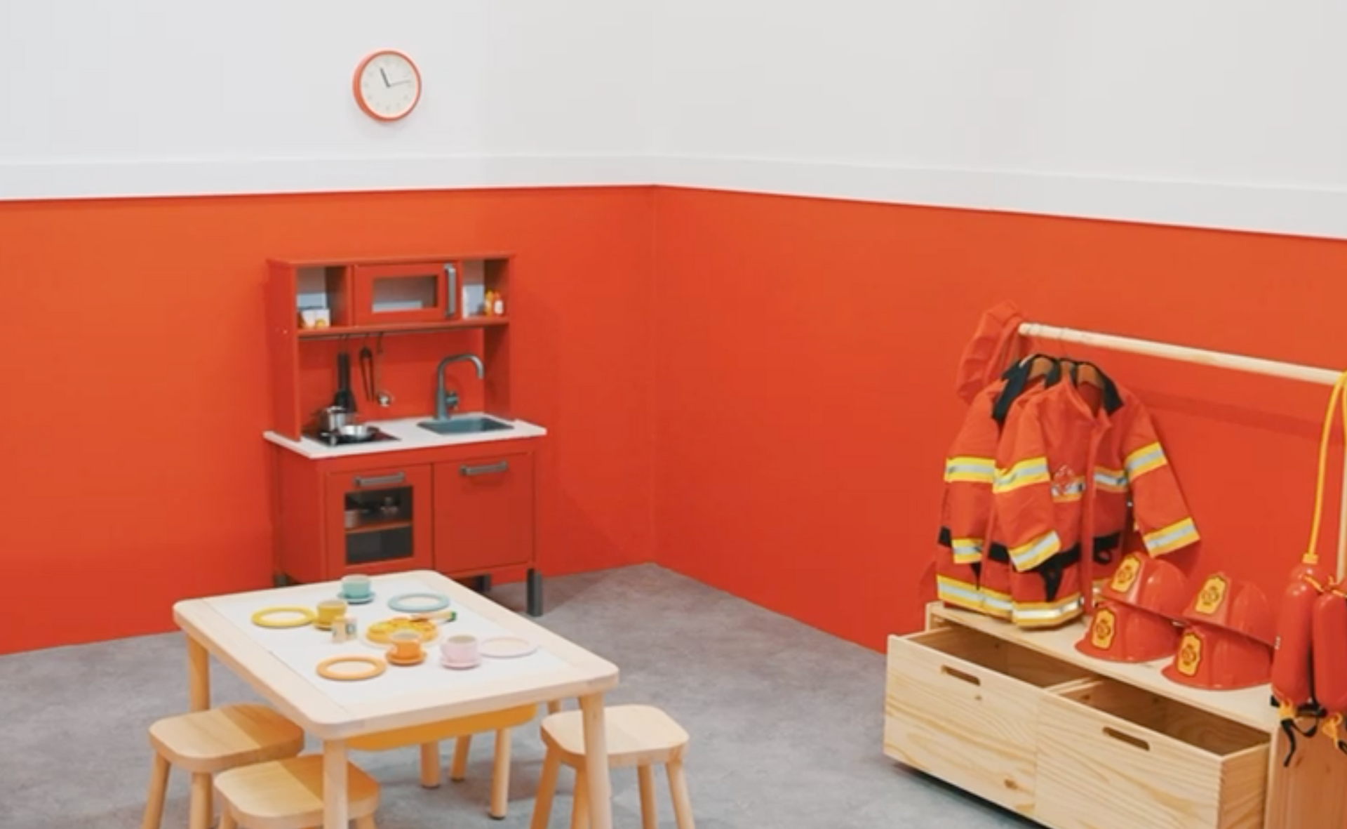 A room with a table , chairs , a clock and a fireman 's uniform.