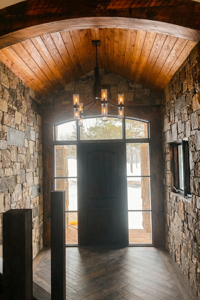 Stone entryway with wooden ceiling and door, a chandelier hangs above the dark door with a glass pane window.