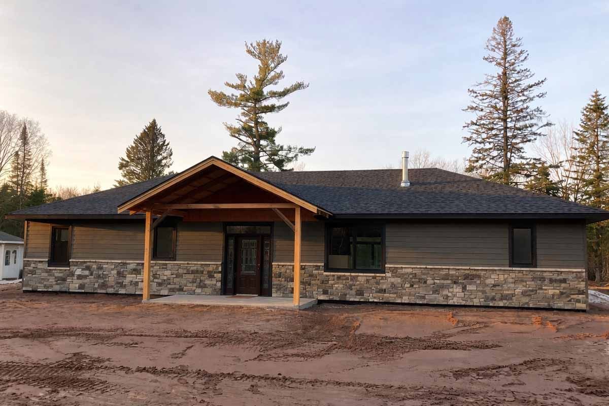 Ranch-style house with stone facade, brown siding, and covered entryway, set amid bare earth and trees.
