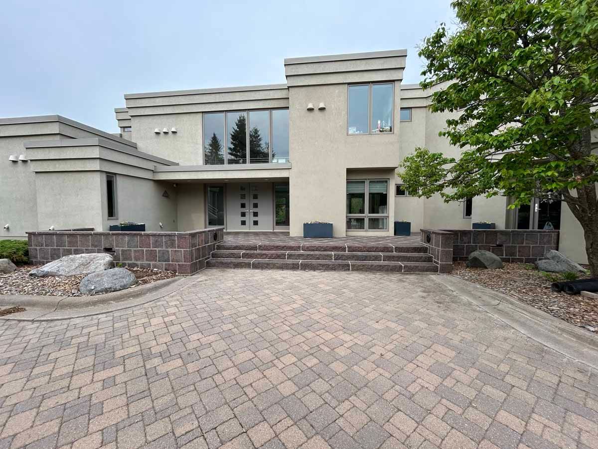 Modern beige home with brick driveway, steps, and large windows.