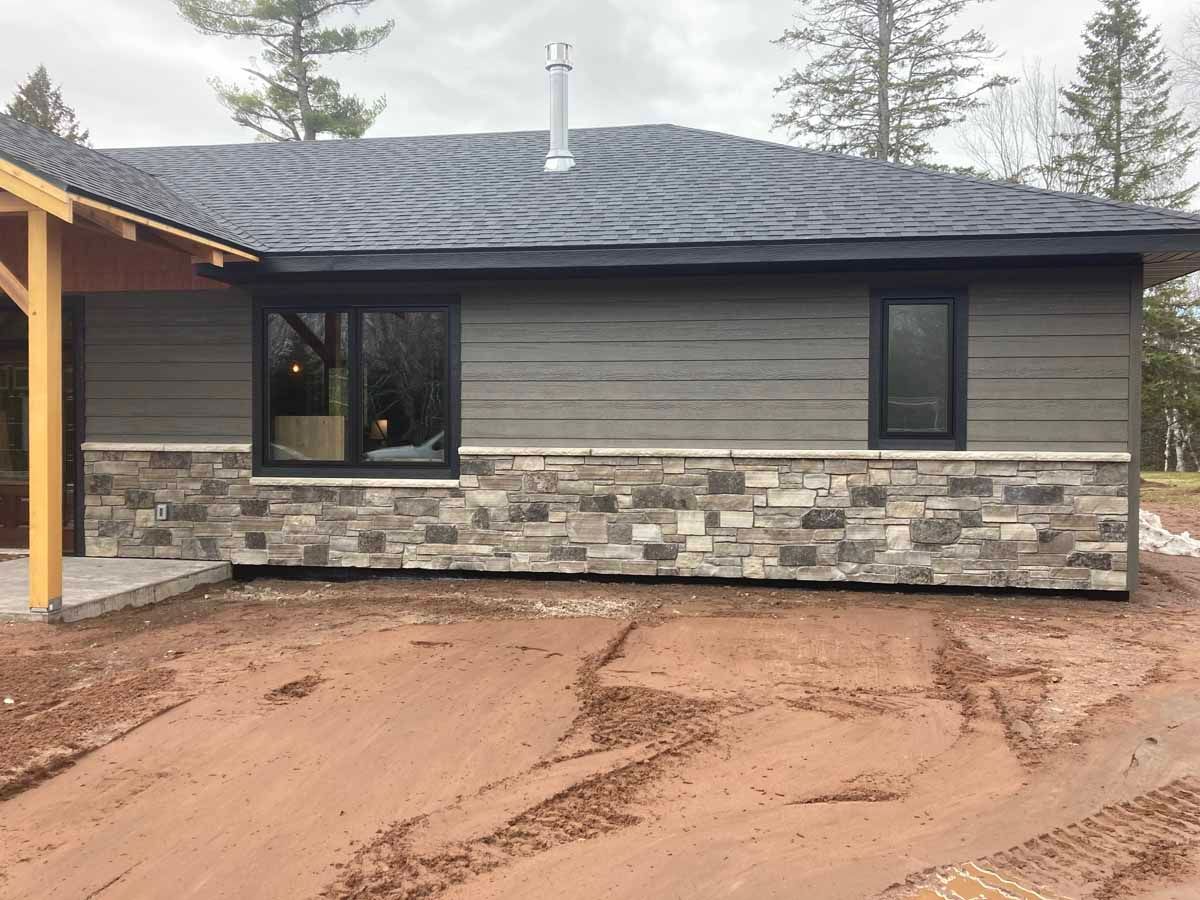 Gray house with stone veneer, dark windows, and a black roof on a muddy lot.