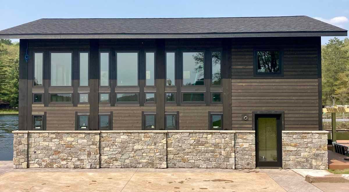Two-story lake house with brown siding, large windows, and a stone wall base. Black roof, clear sky.