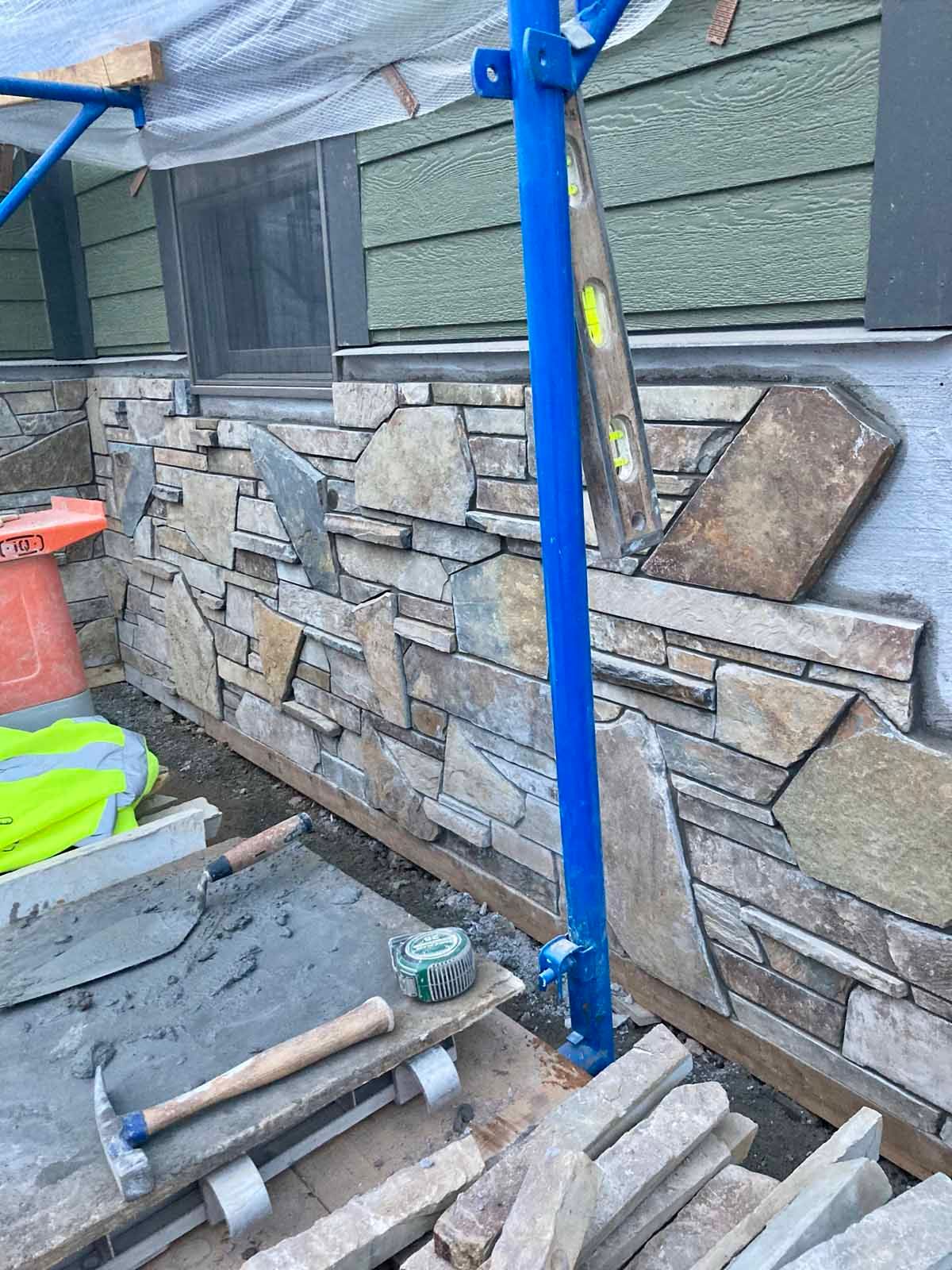 Stone veneer being installed on a building exterior, featuring tan and gray stones, construction tools, and blue scaffolding.