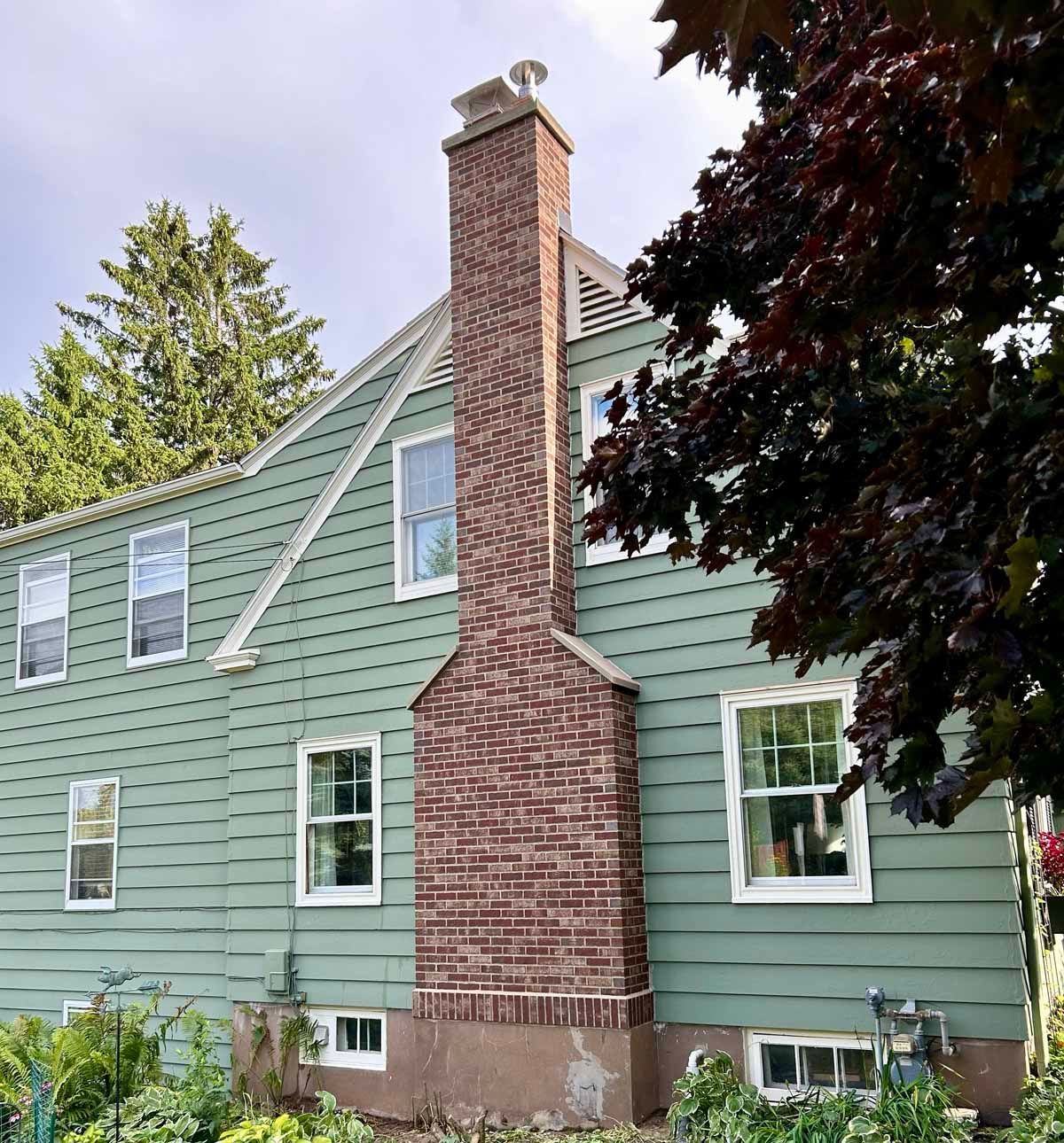 Brick chimney on the side of a green-sided house, tree to the right, cloudy sky.