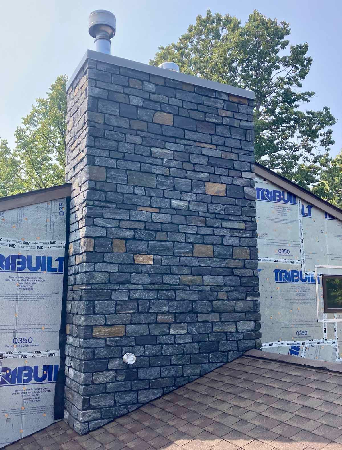 Stone chimney on a house roof, blue-gray stone facade with a metal cap.