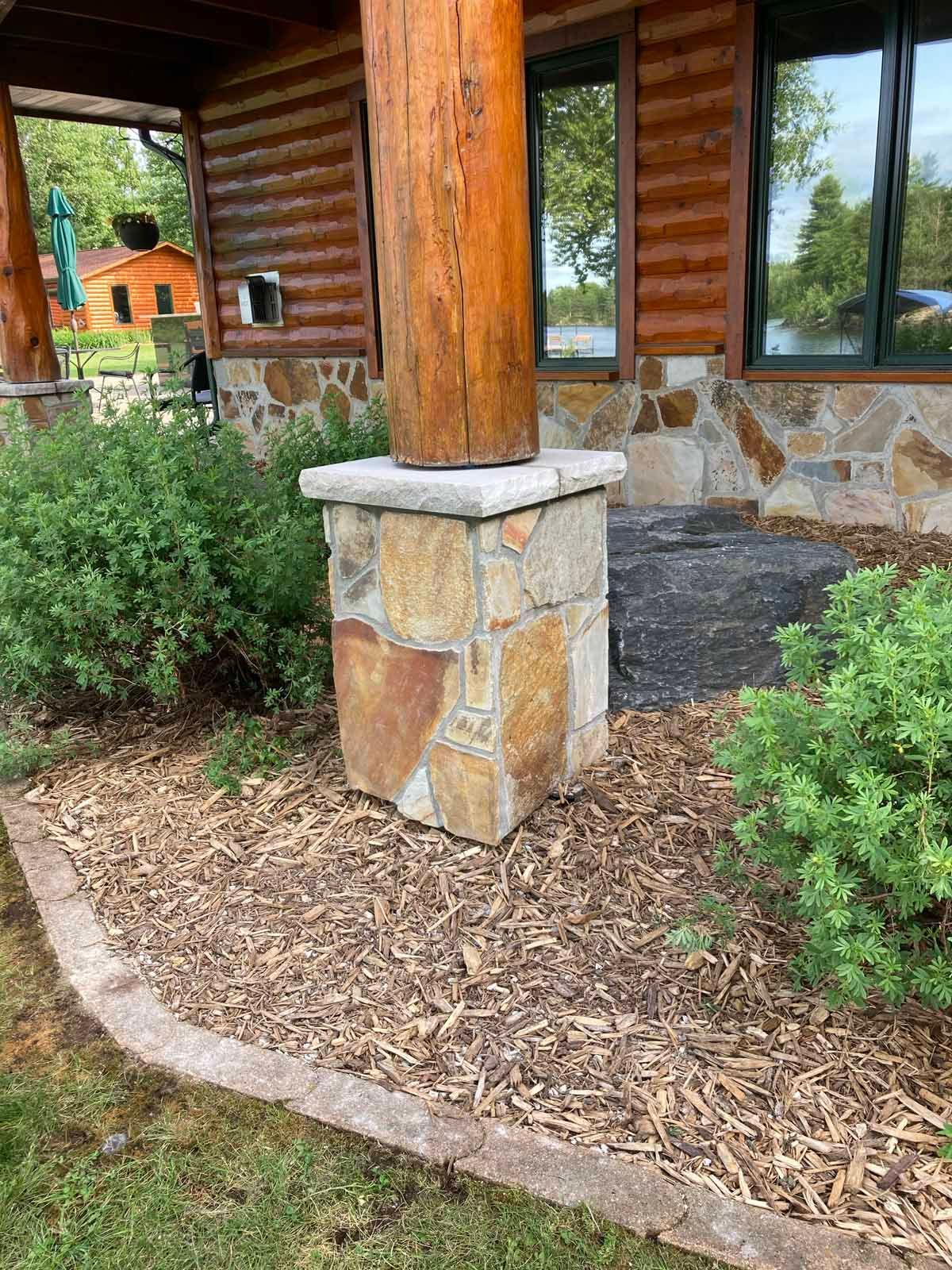 Stone pillar supporting a wooden beam on a porch with stone and wood siding. Landscaping around base.