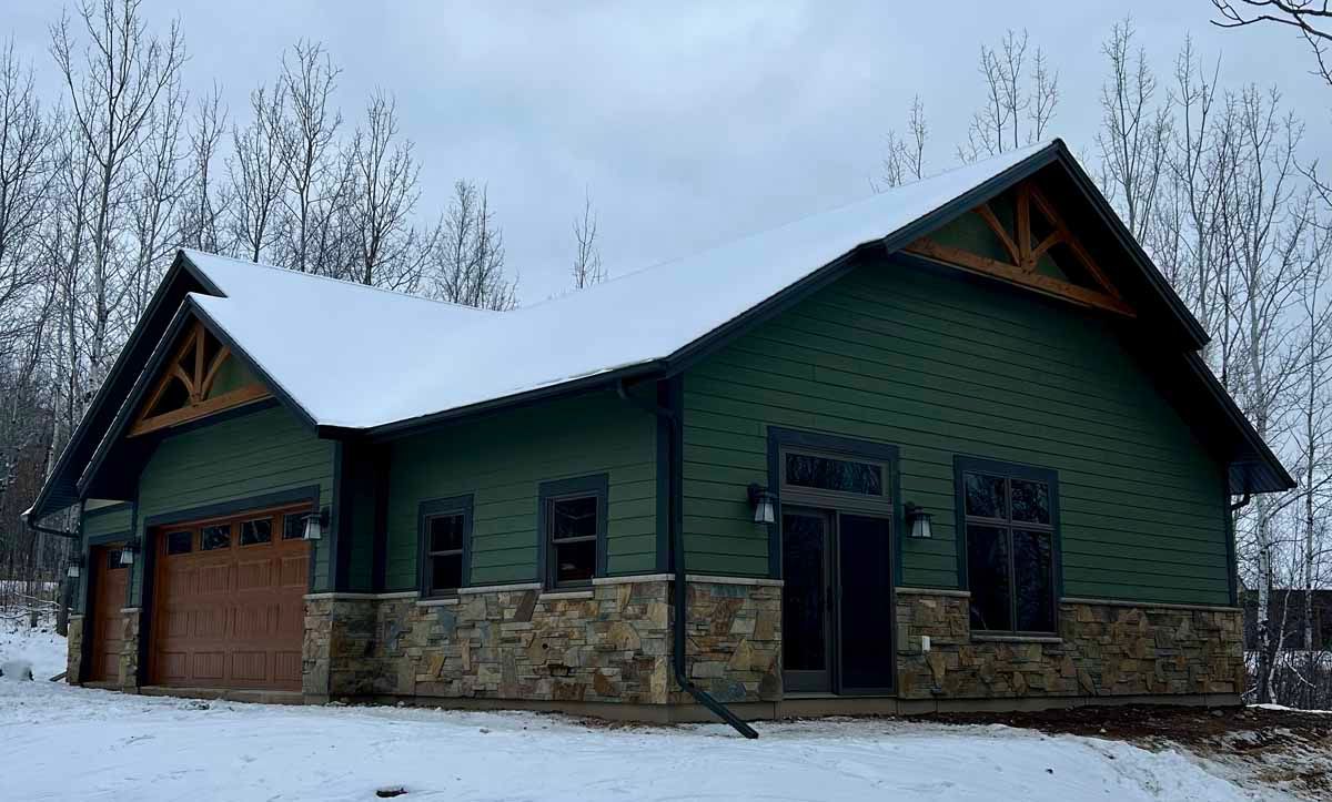 Green building with stone accents, snow-covered roof, brown garage door, and wooden trim in a snowy forest.