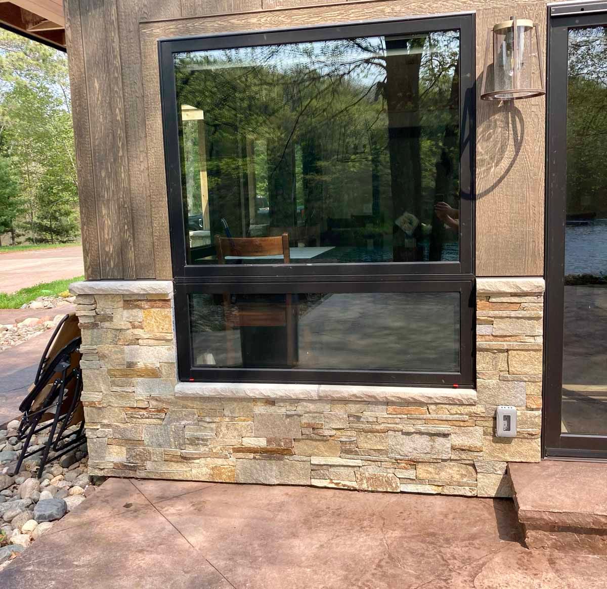 Black-framed window in a stone-covered wall reflecting trees and furniture. Brown stucco and concrete patio.
