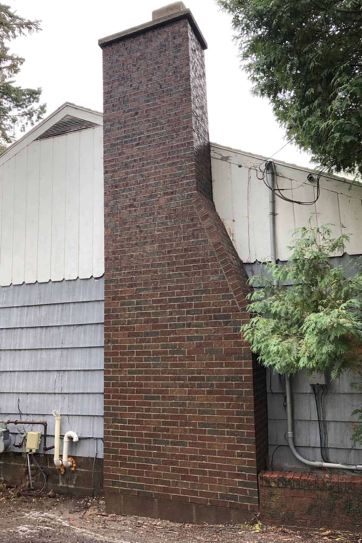 Brick chimney on a house with gray and white siding. Trees and overcast sky in the background.