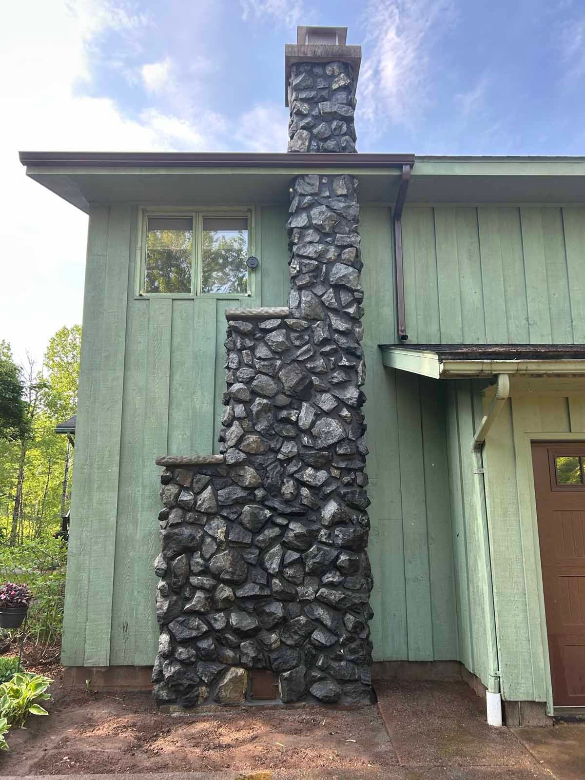 Stone chimney on a green house, with a dark, textured surface against the siding. A window is above the base.