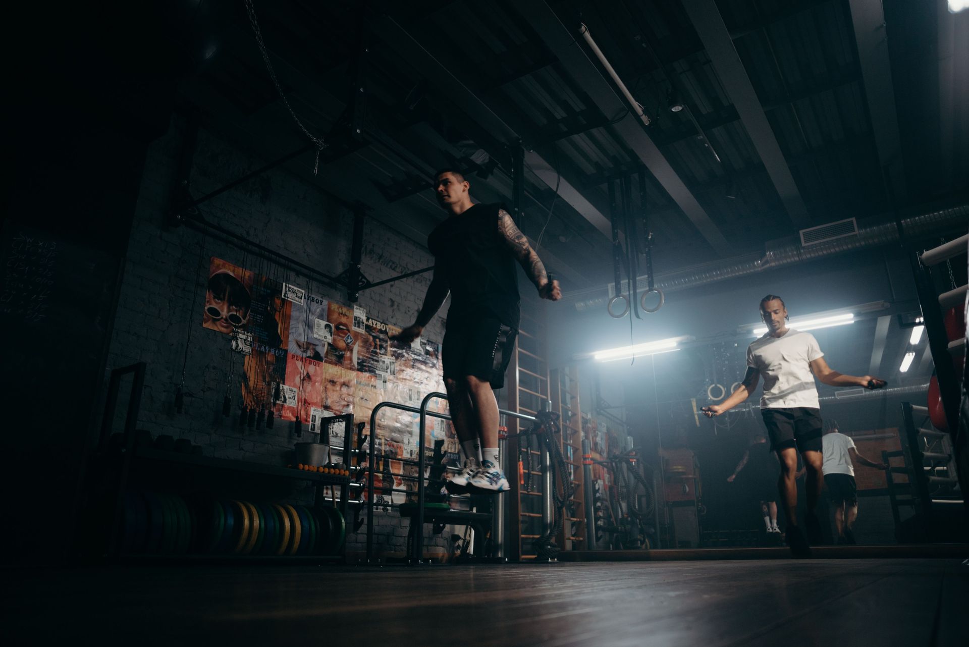 Two people jumping rope in a dim gym with bright backlight and weight racks
