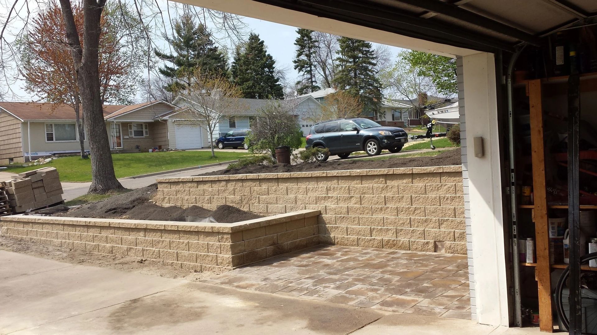 View from a garage of a suburban street, stone retaining wall, lawn, and parked black SUV.