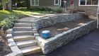 Stone retaining wall and stairs leading to a raised yard beside a house, with a blue bucket on the ledge.
