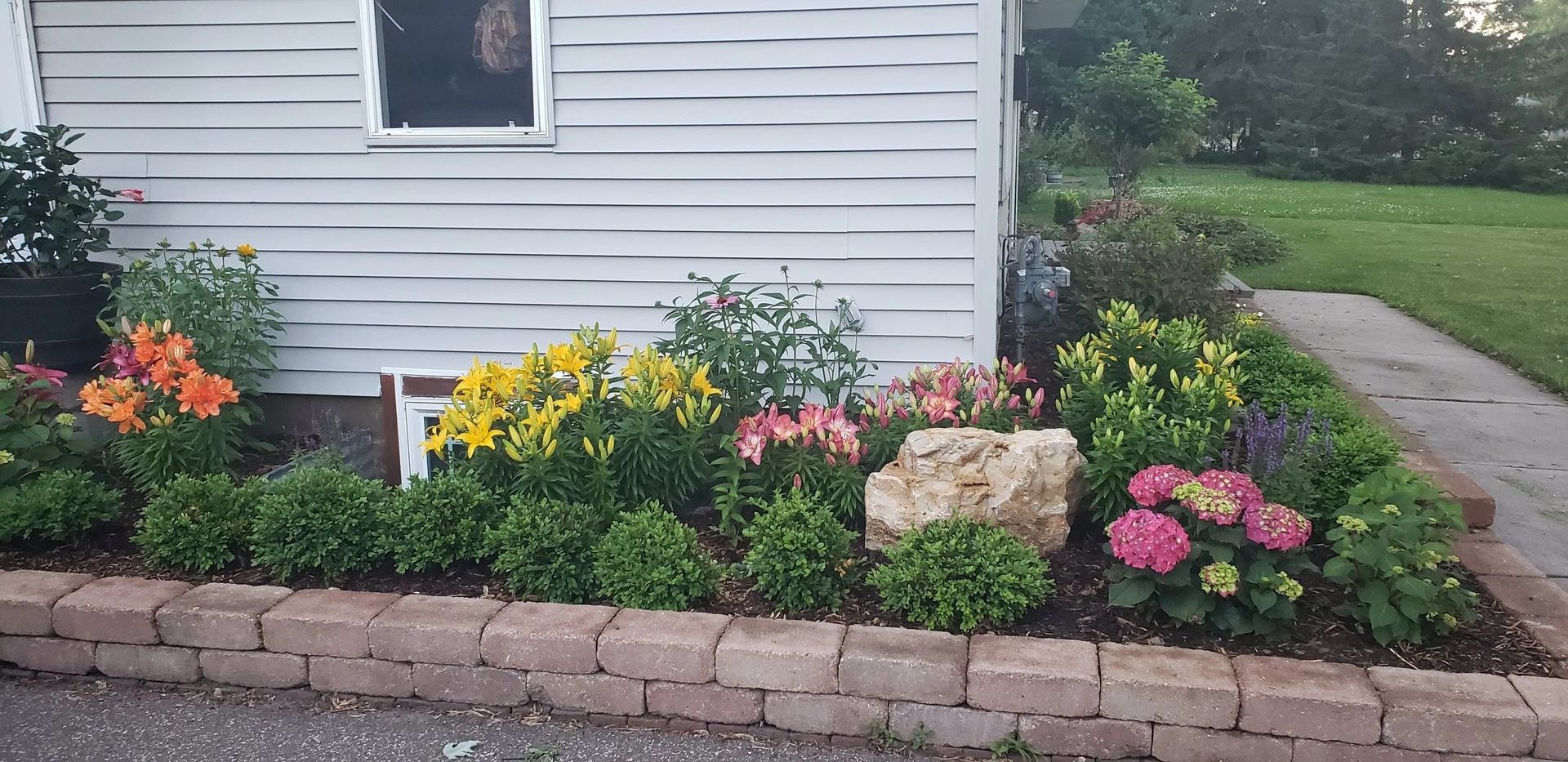 Colorful flower bed with pink, yellow, and orange blooms along a house foundation and stone border