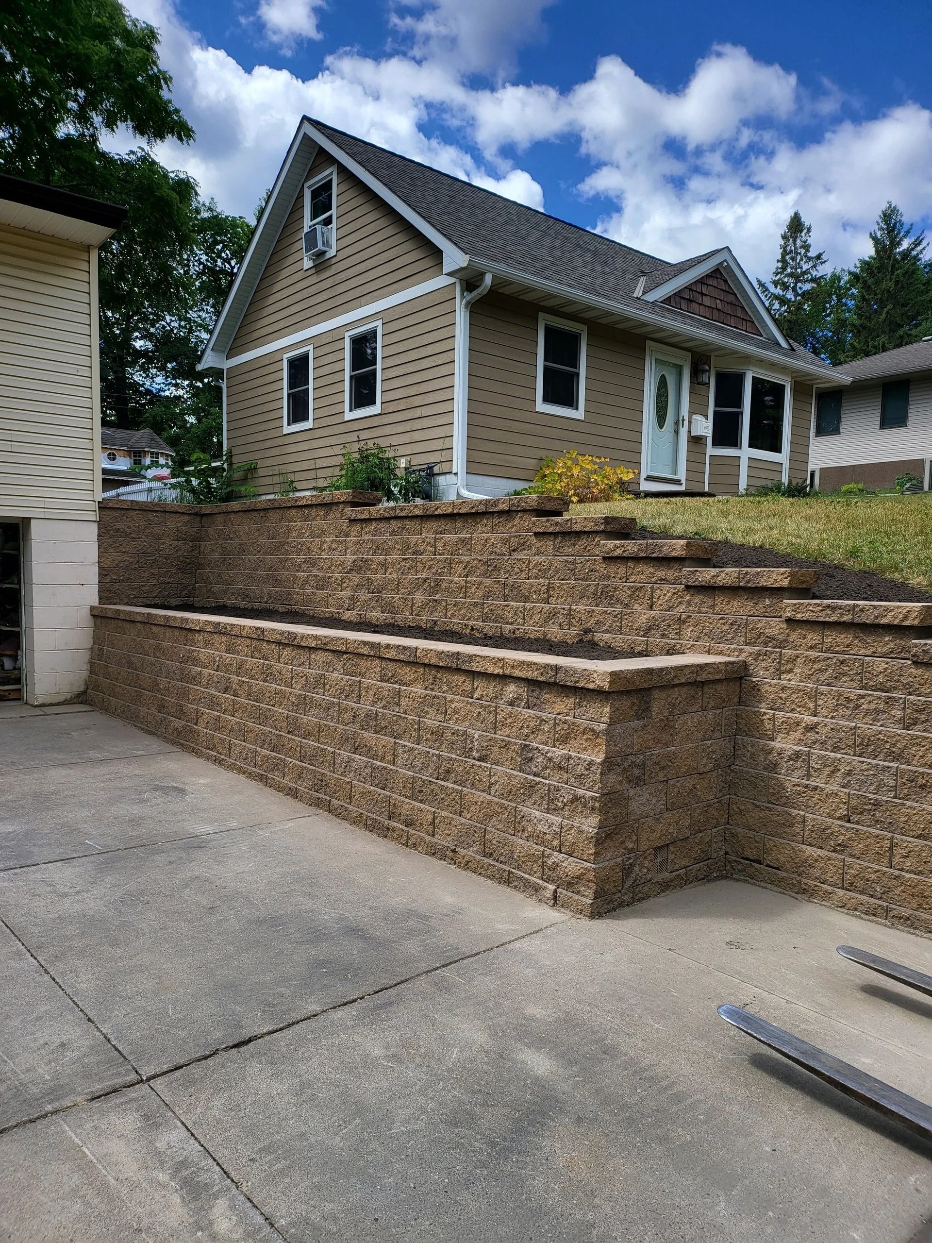 Stone retaining wall beside a tan house with black roof on a sloped driveway under cloudy skies