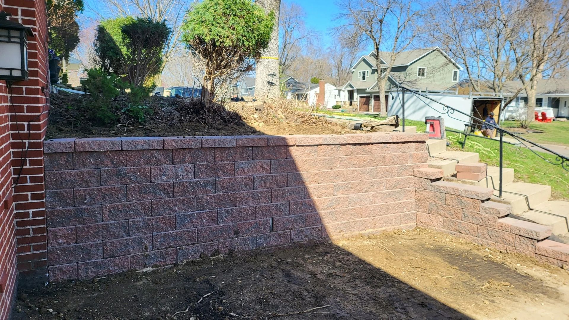 Brick retaining wall and stone steps in a sunny suburban yard with trees and houses in the background