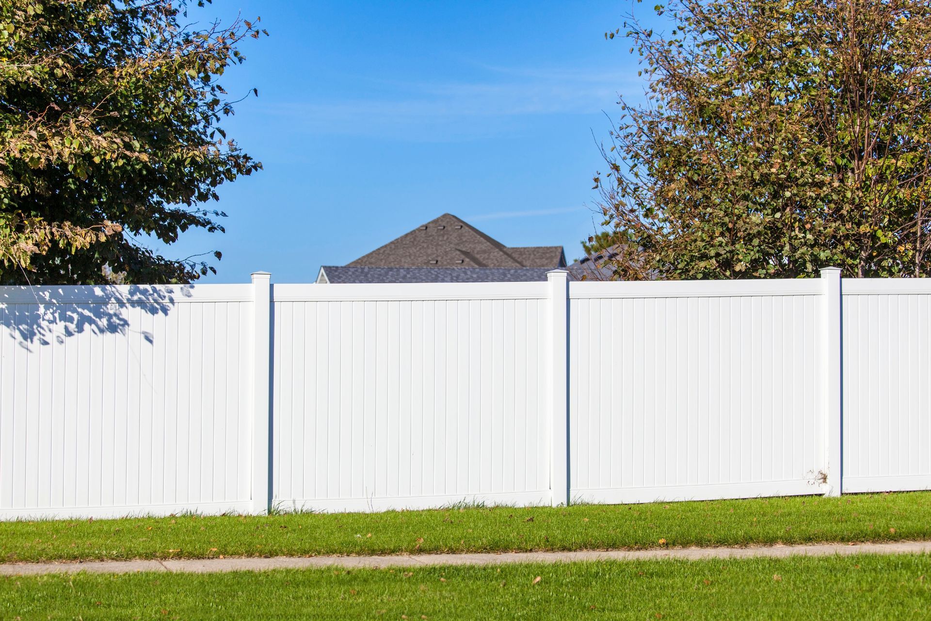 White vinyl privacy fence in a grassy yard with trees and a house roof visible behind it.