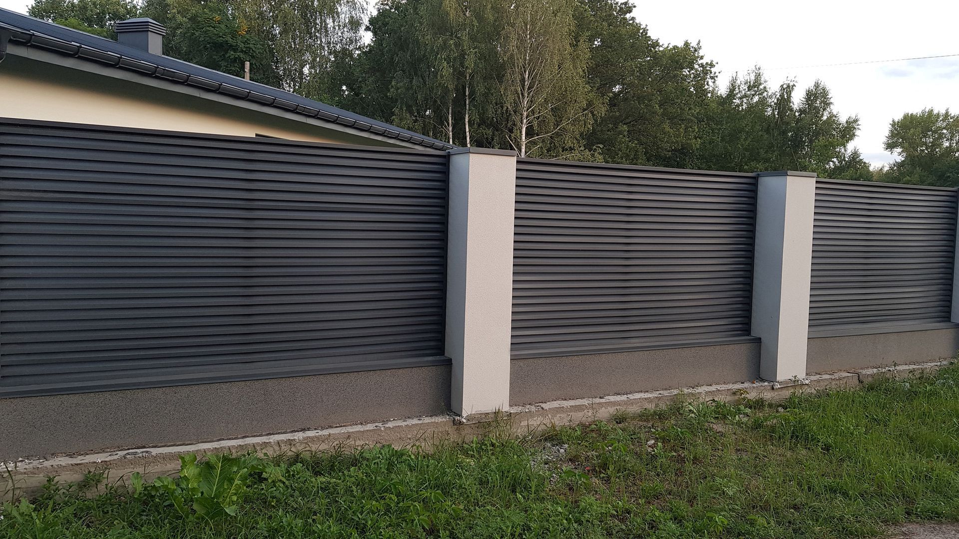 Gray slatted fence with beige pillars along a grassy roadside, with trees in the background