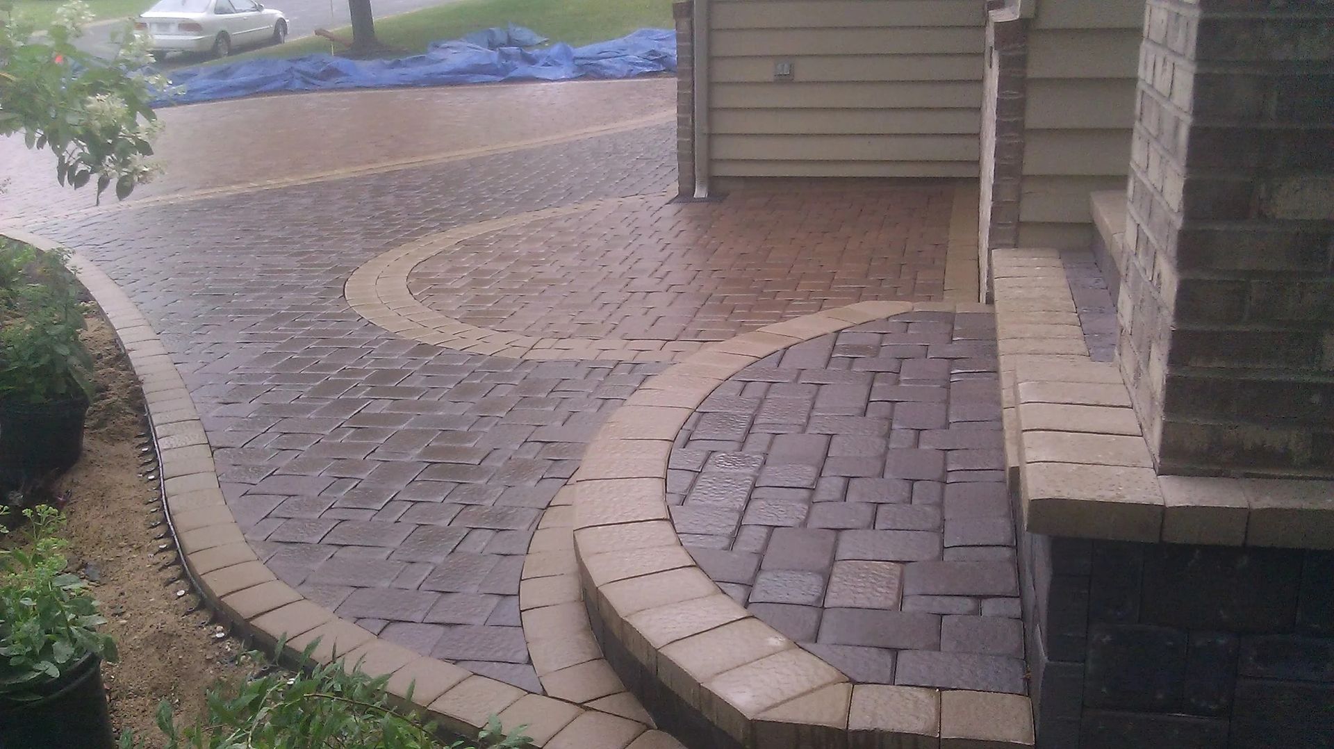Curved paver walkway and patio beside a house, with tan edging and landscaped border.