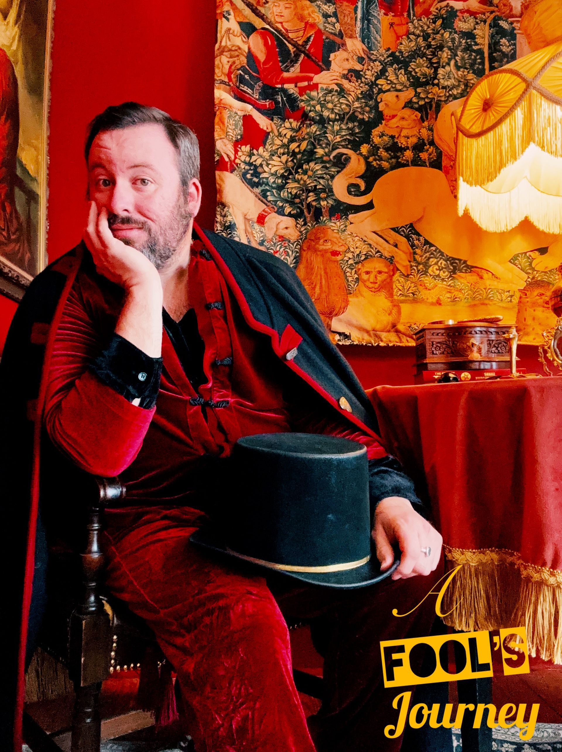 Seated in red velvet with a black top hat, medieval hunting tapestry behind.
