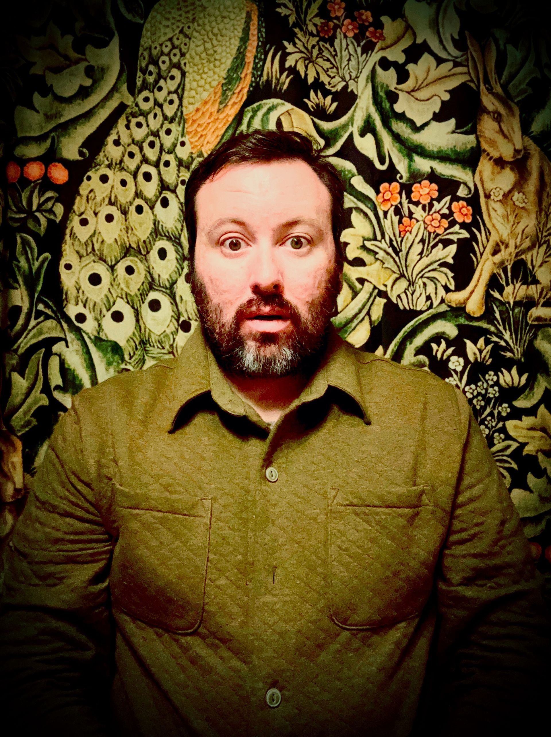 Straight to camera against green William Morris peacock and hare wallpaper.