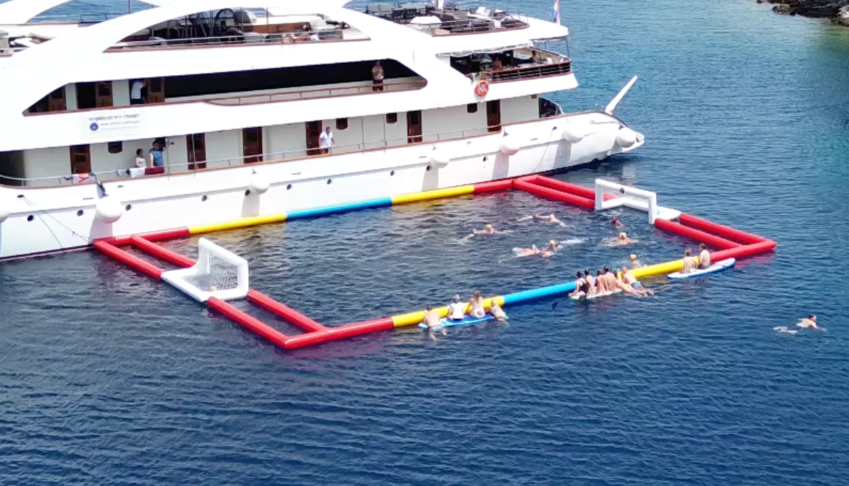 People playing water polo in an inflatable pool attached to a large white yacht in blue water.