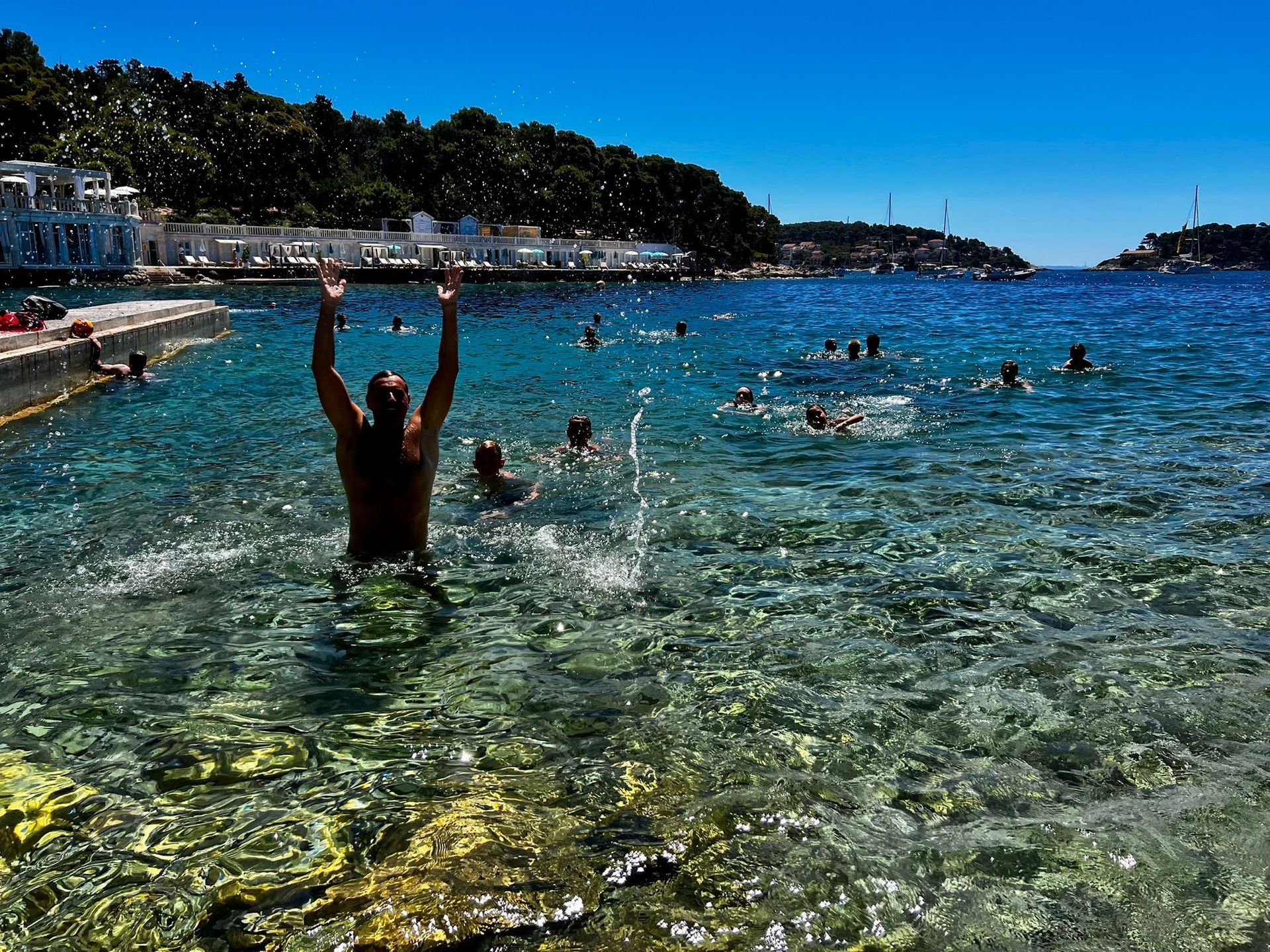 Man in clear water with arms raised, enjoying a swim with other people in a bay with buildings and trees in the background.