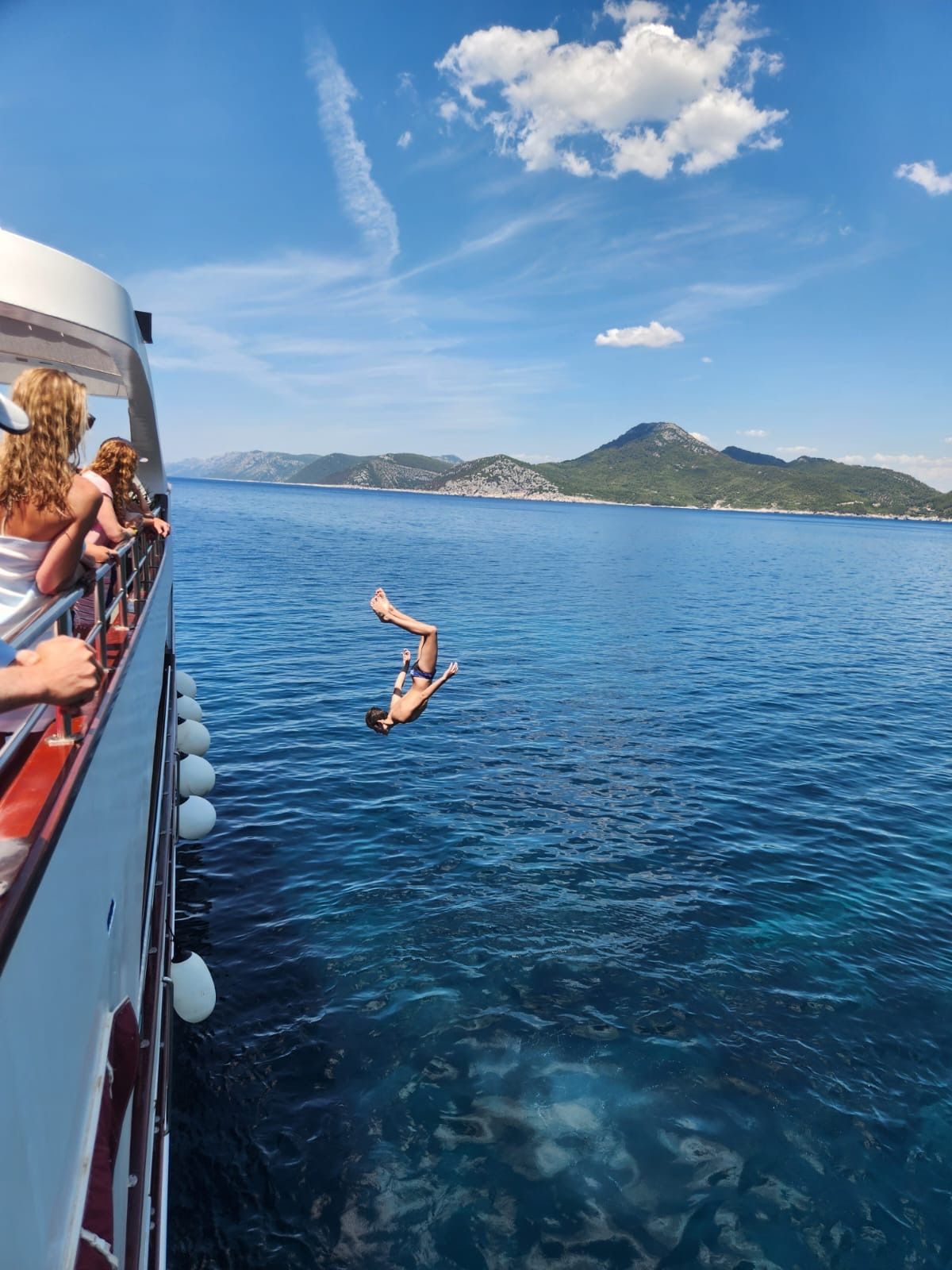 Person dives from a boat into the blue sea. Island and blue sky in background.