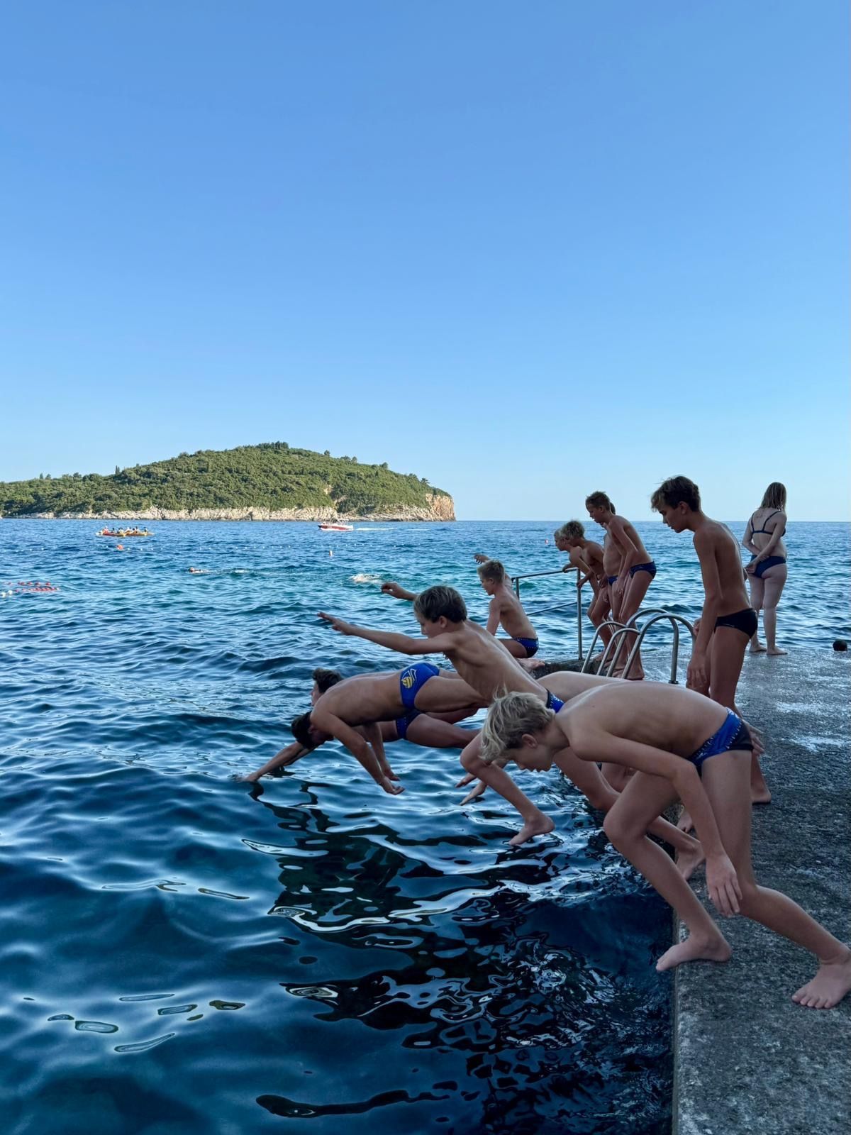 People jumping into the sea from a concrete dock on a sunny day with an island in the distance.